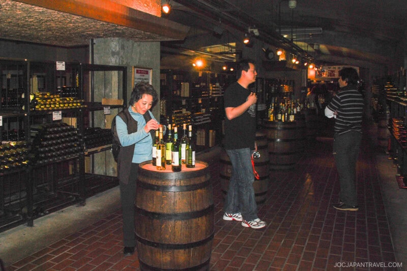 People sample wine at wooden barrels inside a dimly lit Yamanashi wine cellar, surrounded by shelves filled with wine bottles. Three individuals are seen tasting or examining bottles.