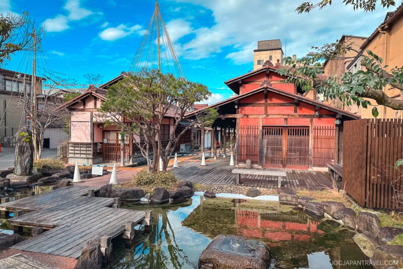 A traditional Japanese building with wooden decks and a small pond in the foreground, surrounded by trees and rocks under a bright blue sky, evokes the tranquil ambiance of Yamashiro Onsen.