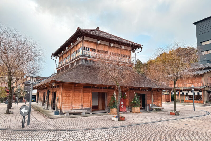 A traditional Japanese wooden building with a tiled roof sits on a paved street in Yamashiro Onsen. Some trees without leaves and potted plants surround the structure beneath the partly cloudy sky.
