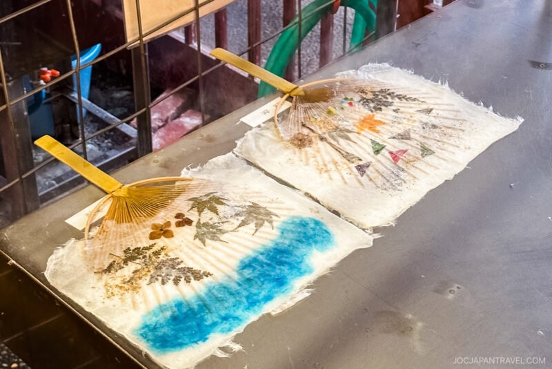 Two handmade Japanese paper fans from Yunokuni no Mori, each with wooden handles, are decorated with pressed flowers and leaves. One features blue coloring at the bottom, while both display natural plant designs on a textured surface, drying on a metal table.
