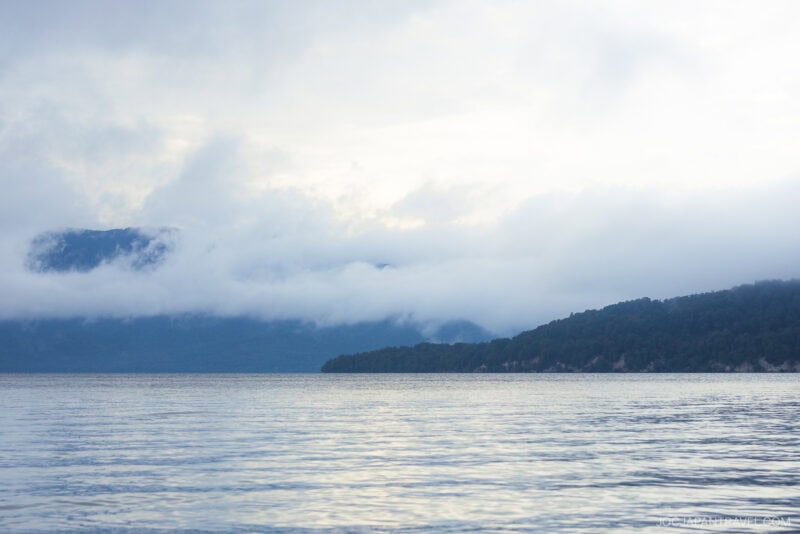 A calm Lake Kussharo with gentle ripples in the foreground, a forested hill in the background, and low clouds partially covering distant mountains under a cloudy sky.