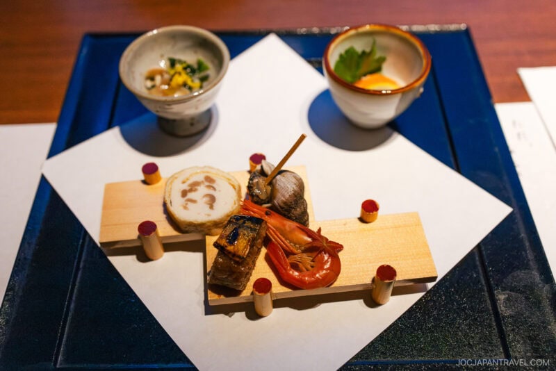 A beautifully arranged Japanese kaiseki appetizer platter, inspired by the tranquil waters of Lake Kussharo, features assorted seafood, vegetables, and sliced roll on small wooden boards, flanked by two garnish-filled bowls on a white paper-lined tray.
