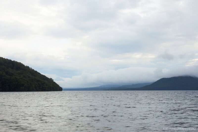 A calm Lake Kussharo stretches into the distance under a cloudy sky, with forested hills on the left and right sides meeting at the horizon. The water appears gentle and slightly rippled.