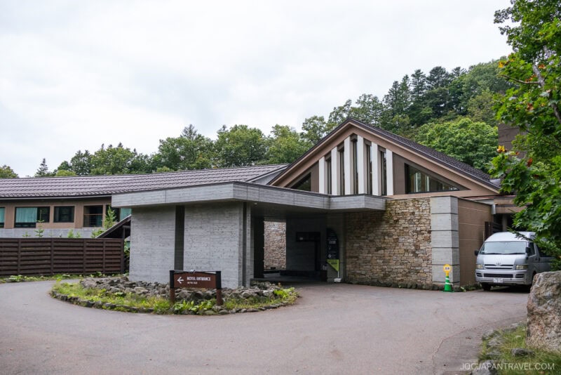 Modern hotel entrance with stone walls, sloped roof, and large windows surrounded by trees near Lake Kussharo; a small sign says Hotel Okunoyu and a white van is parked by the covered driveway.