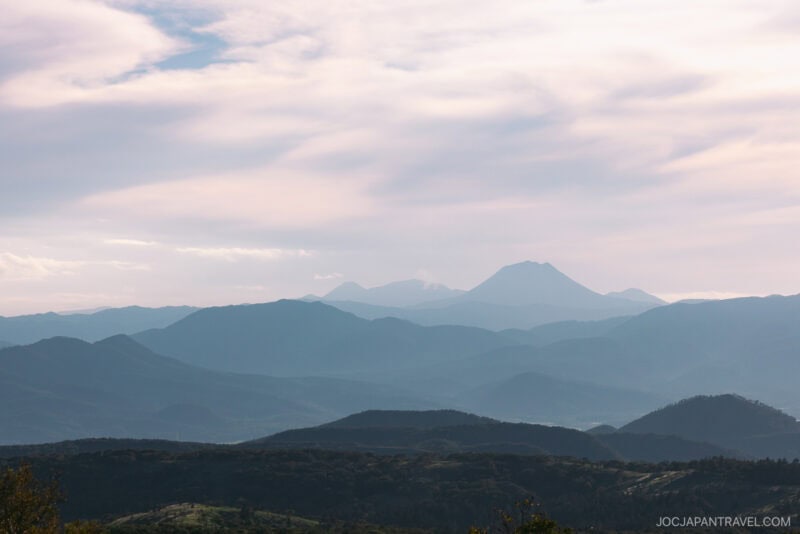 Mountain ranges under a soft, cloudy sky overlook Lake Kussharo, with layers of blue and green hills fading into the distance, creating a peaceful and atmospheric landscape.