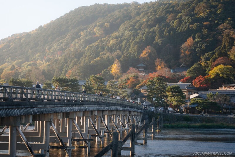 A long wooden bridge crosses a river in Arashiyama autumn, with people walking on it, surrounded by colorful trees and hills in the background under soft sunlight.