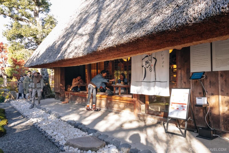People gather outside a traditional Japanese building with a thatched roof in Arashiyama autumn, surrounded by a serene garden. Some stand, while others sit by open windows beneath a white curtain with artwork. Sunlight brightens the peaceful scene.