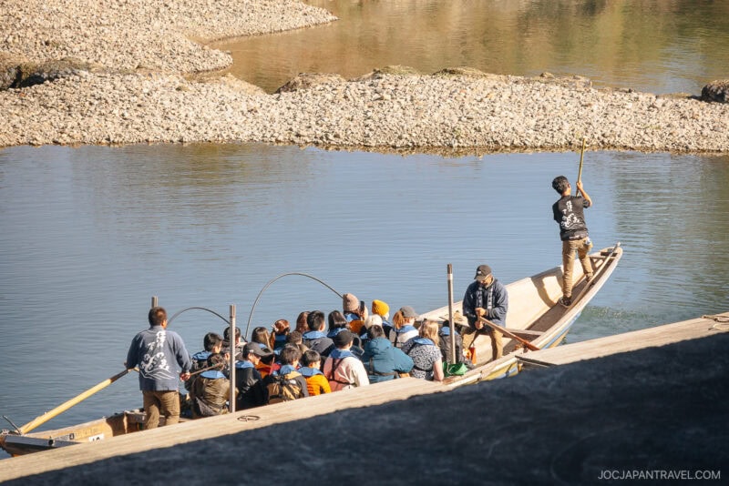A group of people sit in a wooden boat steered by two guides with poles, floating on a calm river near a rocky shore under bright sunlight—an idyllic Arashiyama autumn scene.
