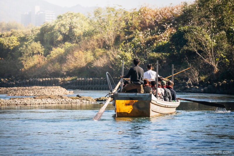 A group of people ride in a wooden boat on a calm river in Arashiyama, surrounded by lush autumn greenery and trees, with sunlight illuminating the scene from above.