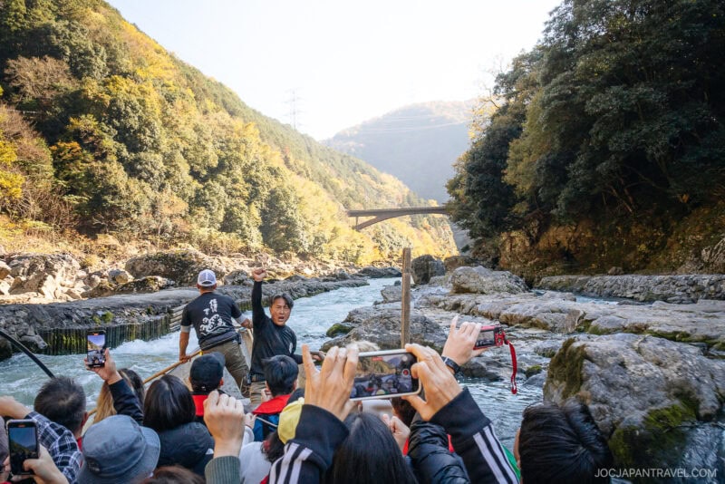 A group of tourists in a wooden boat tour the scenic Arashiyama river in autumn, surrounded by rocky banks and forested hills, with many passengers taking photos of the vibrant landscape ahead.
