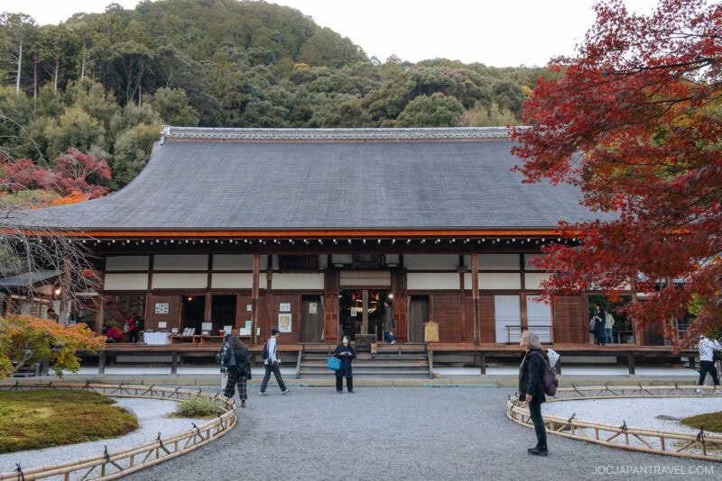 Visitors walk in front of a traditional Japanese wooden temple surrounded by trees with vibrant Arashiyama autumn foliage, set against a backdrop of forested hills.