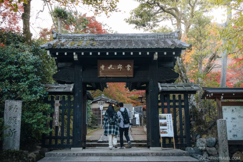 Two people walk through a traditional Japanese wooden gate in Arashiyama, surrounded by trees with vibrant autumn leaves. Signs and stone pillars mark the entrance to this peaceful temple or garden area.