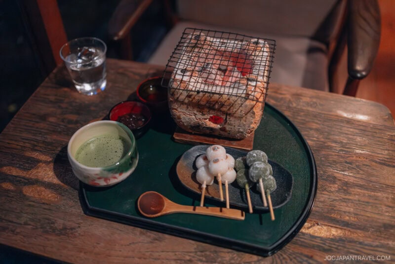 A wooden table set with matcha tea, skewered mochi balls, dipping sauces, and a small charcoal grill evokes the cozy charm of Arashiyama autumn, with a glass of water and a wooden chair in the background.