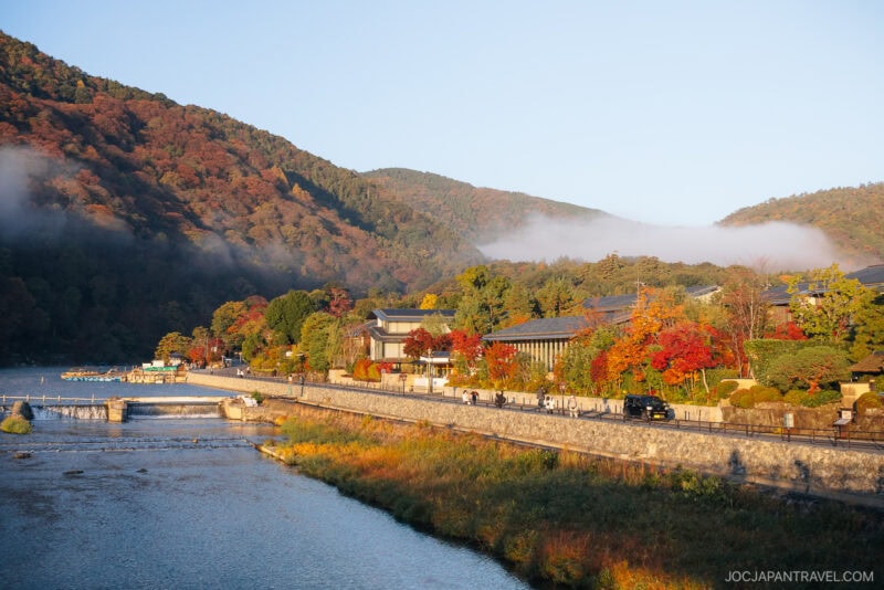 A scenic Arashiyama autumn riverside view with colorful foliage, traditional and modern buildings, cars on a road, and tree-covered hills under a clear sky with low mist in the background.