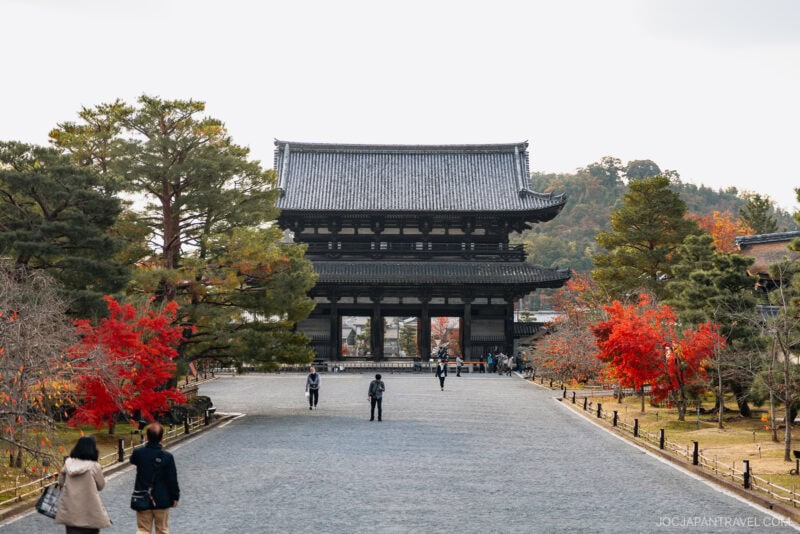 A wide stone path leads to a large traditional Japanese temple gate, surrounded by trees with vibrant Kyoto autumn foliage and people walking on the pathway.