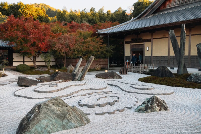 A Japanese Zen garden with carefully raked gravel, large rocks, and upright stone slabs near a traditional wooden building, surrounded by Kyoto autumn foliage in vibrant colors.