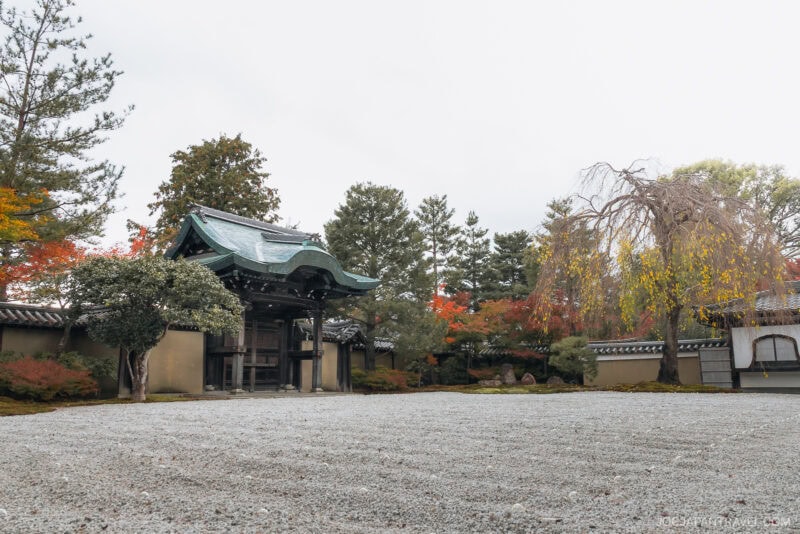 A traditional Japanese gate stands in a gravel courtyard, surrounded by trees with Kyoto autumn foliage in shades of green, yellow, and red. The overcast sky adds to the calm atmosphere of this serene garden scene.
