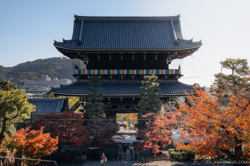 A traditional Japanese temple gate with a tiled roof stands amid vibrant Kyoto autumn foliage, as people stroll beneath it. Hills and a clear sky form the scenic background.