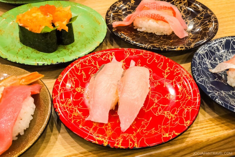 Several colorful plates of sushi, including nigiri with raw fish and gunkan with orange roe and sea urchin, are arranged on a wooden table, evoking the lively variety found at a conveyor belt sushi restaurant.