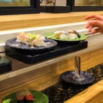 How to Enjoy Conveyor Belt Sushi in Japan: A Complete Guide A hand reaches for a plate of sushi from a conveyor belt sushi restaurant. Several plates with sushi pieces glide by on the moving black track, offering a tempting selection.