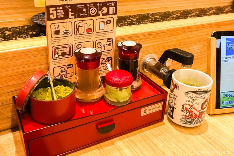Condiments, green tea powder, and a faucet for hot water are arranged on a wooden counter. A cup with green tea sits beside illustrated tea instructions at a conveyor belt sushi restaurant in Japan.