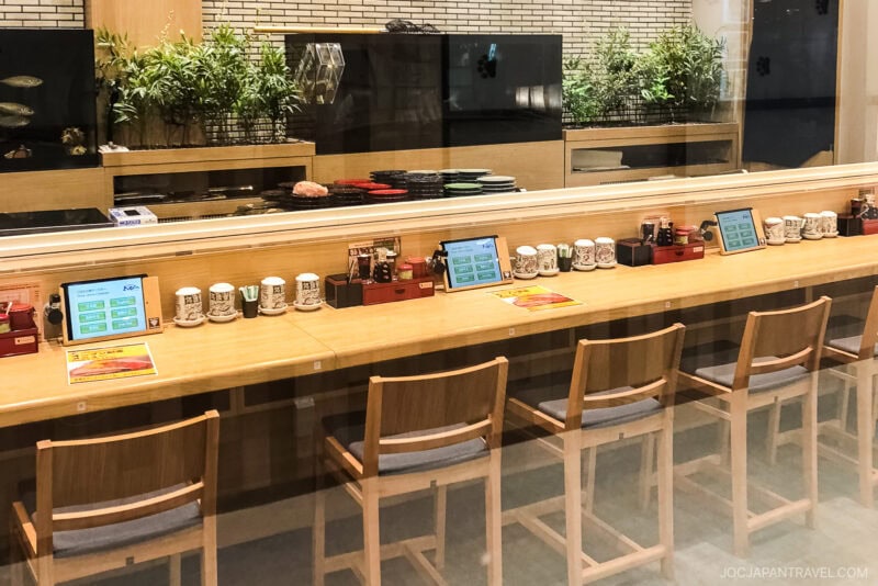 A row of wooden chairs faces a conveyor belt sushi bar with a glass divider, each seat equipped with a touchscreen, cups, and condiments. Green plants and sushi platters glide along the counter behind the glass.