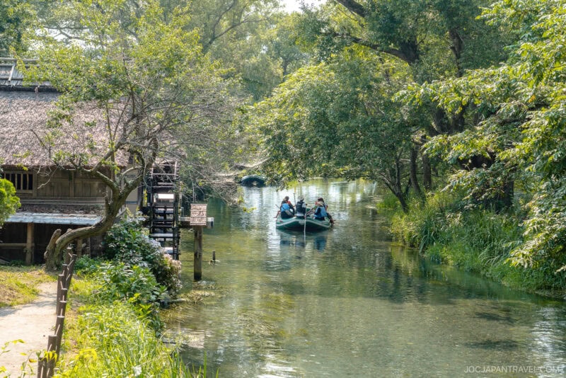 A small group of people ride a boat down a clear, tree-lined canal at the Daio Wasabi Farm, passing a rustic wooden building with a thatched roof, surrounded by lush greenery on a sunny day.