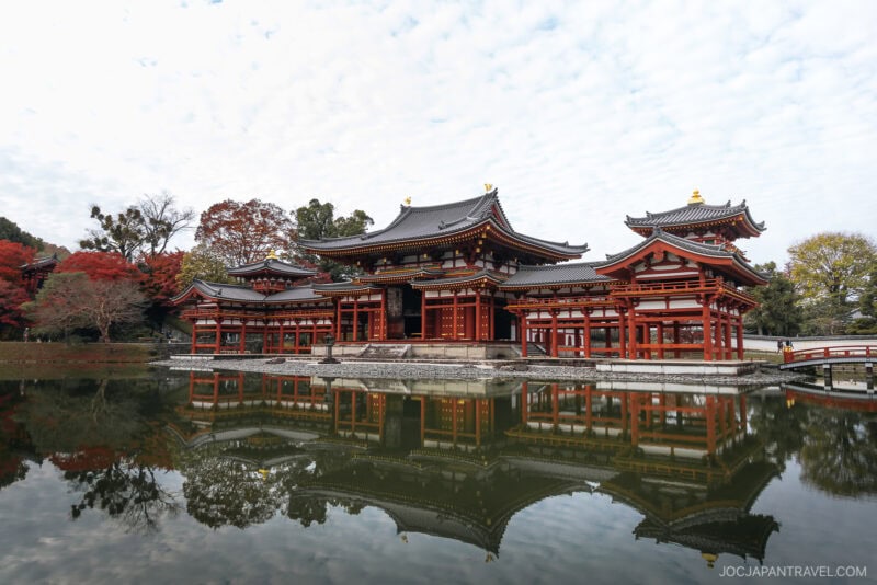 A traditional Japanese temple in Uji with intricate red and white architecture is reflected in a calm pond, surrounded by autumn trees with red and green foliage under a partly cloudy sky.