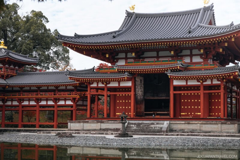A traditional Japanese temple in Uji with ornate red and white wooden structures, curved tiled roofs, and gold accents, reflected in a calm pond with a stone lantern in front.