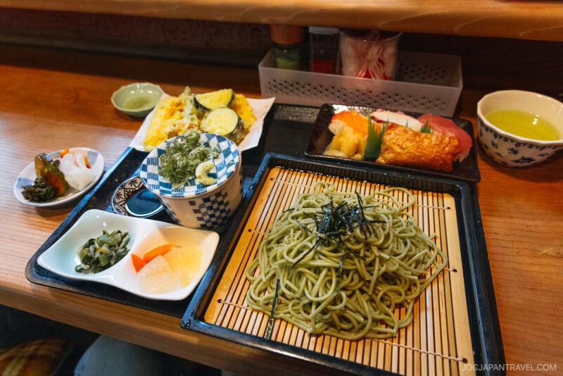 A Japanese meal set on a wooden table features Uji green tea, cold green soba noodles, tempura vegetables, sushi, pickled vegetables, seaweed, and dipping sauce. The dishes are arranged neatly on trays and plates.