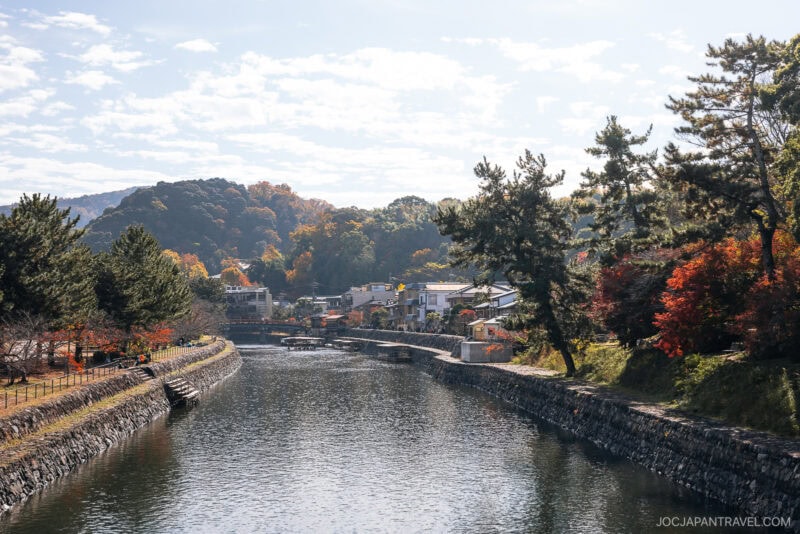 A serene river flows through the quiet town of Uji, bordered by stone walls, trees with autumn foliage, and distant hills under a partly cloudy sky. Some buildings are visible along the riverbank.