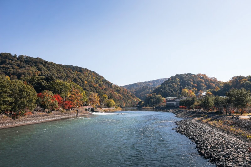 A wide river, reminiscent of the scenic Uji region, flows between tree-covered hills with autumn foliage under a clear blue sky. Rocky banks line the water, and a few buildings are visible among the trees on the right.