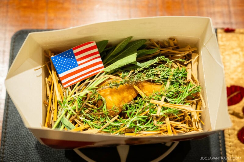 A cardboard box from Den restaurant contains a pastry topped with herbs and straw, next to an American flag toothpick. The box rests on a table with a patterned cloth partly visible.