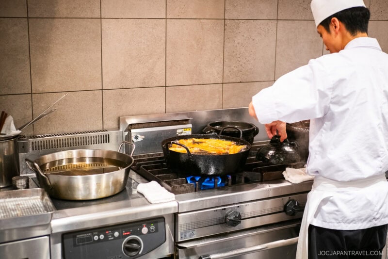 A chef in a white uniform and hat cooks at a gas stove in a commercial kitchen at Den restaurant, tending to a steaming pot of food amid an array of cooking utensils and equipment.