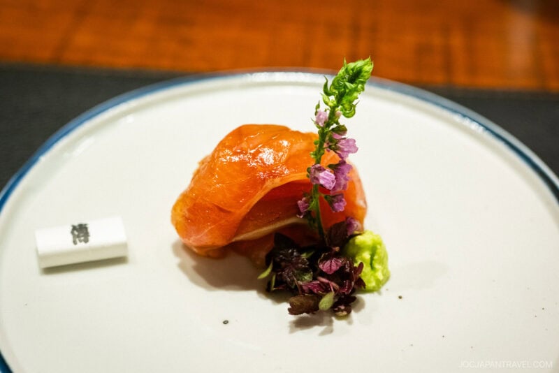 A piece of delicate salmon sashimi is artfully plated with wasabi, microgreens, and a sprig of purple flowers on a white plate with a blue rim at Den Restaurant. A rolled napkin or paper rests beside the fish.