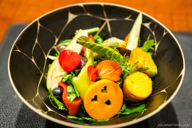 A colorful salad in a black bowl at Den restaurant features sliced persimmon with heart-shaped cutouts, cherry tomatoes, lettuce, potatoes, leafy greens, edible flowers, and other fresh vegetables. The background is softly blurred.