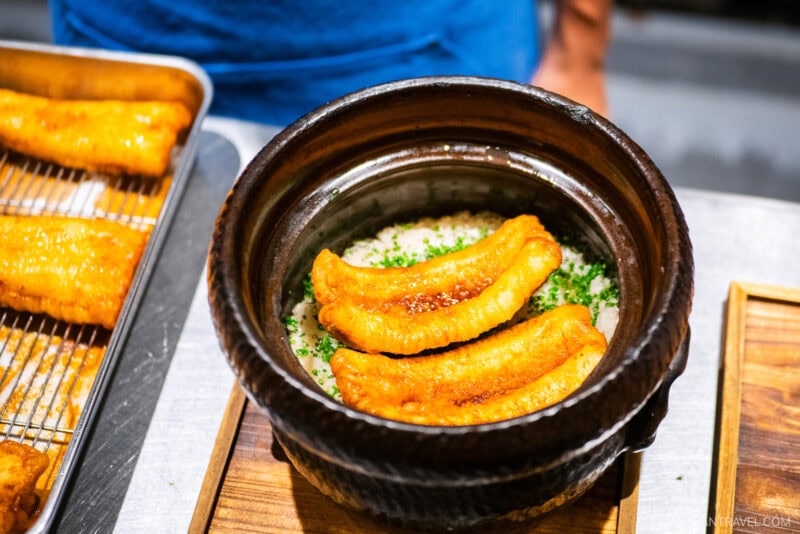 A close-up of a clay pot at Den restaurant, filled with rice and topped with three golden-brown fried eel fillets, garnished with chopped green herbs. More fried eel pieces are visible on a tray to the side.