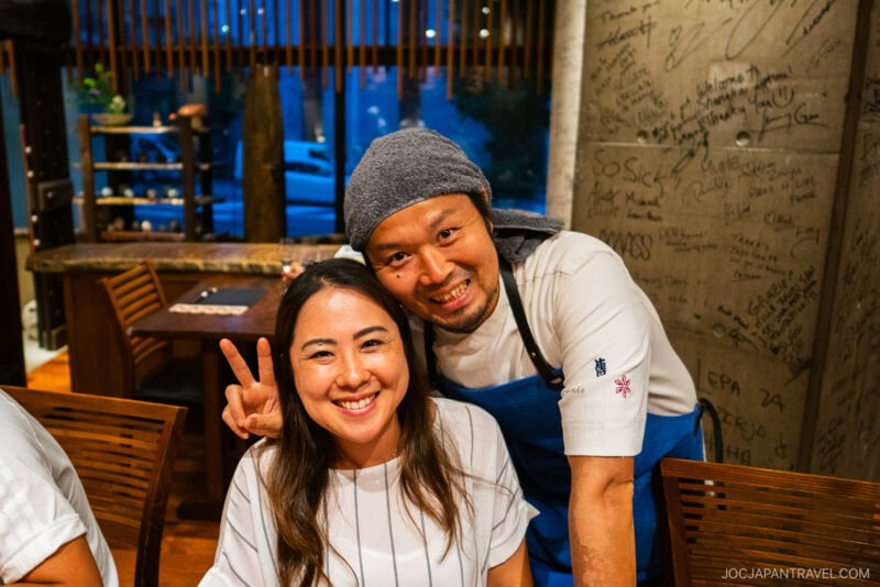 A smiling woman sits at a table in Den restaurant while a chef in a blue apron and gray headscarf stands beside her, making a peace sign behind her head. The restaurant wall behind them is covered in handwritten messages.