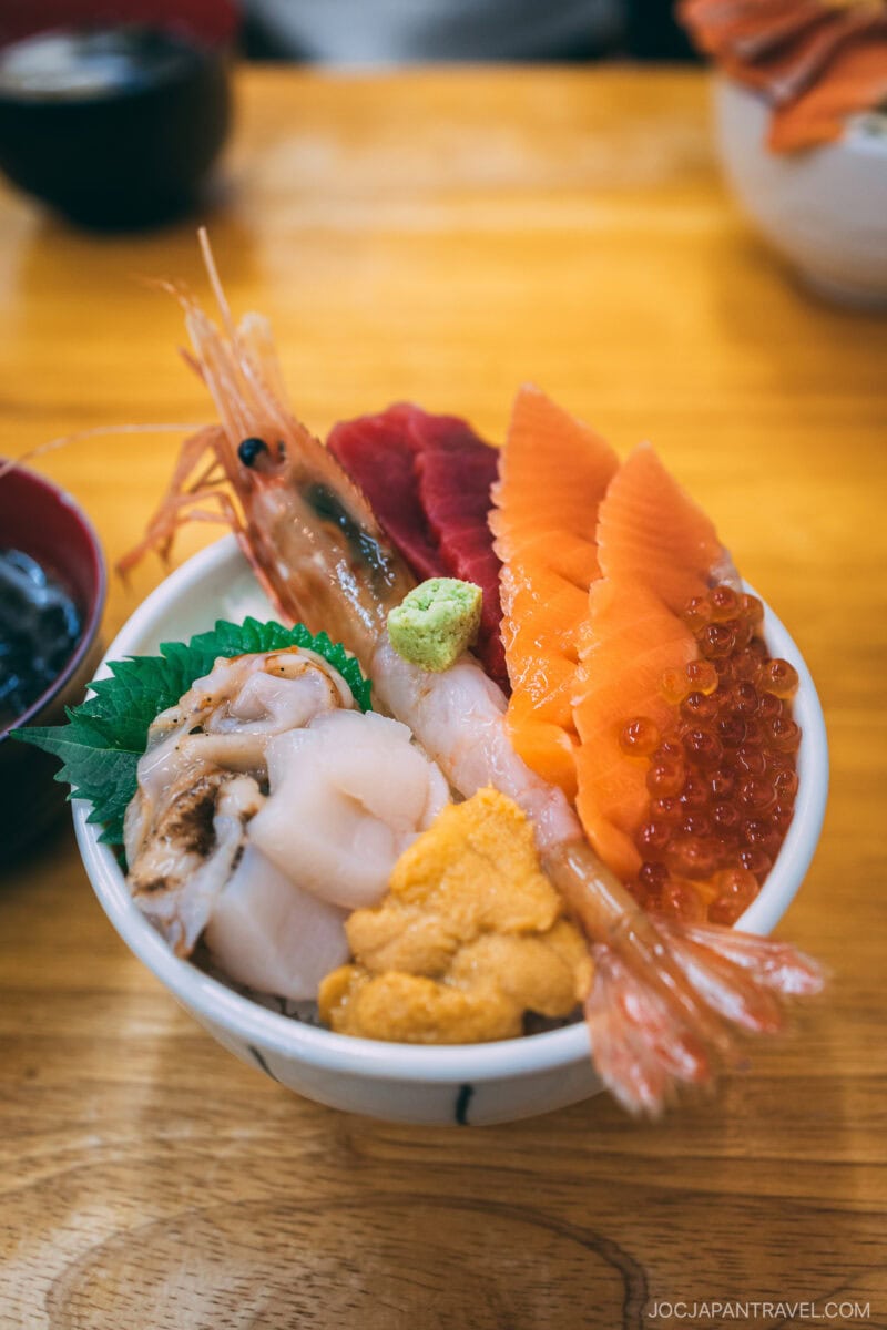 A bowl of assorted sashimi donburi—shrimp, salmon, tuna, scallops, uni, salmon roe, and wasabi on rice—served in a white bowl on a wooden table; a delicious highlight from any Hokkaido food guide.