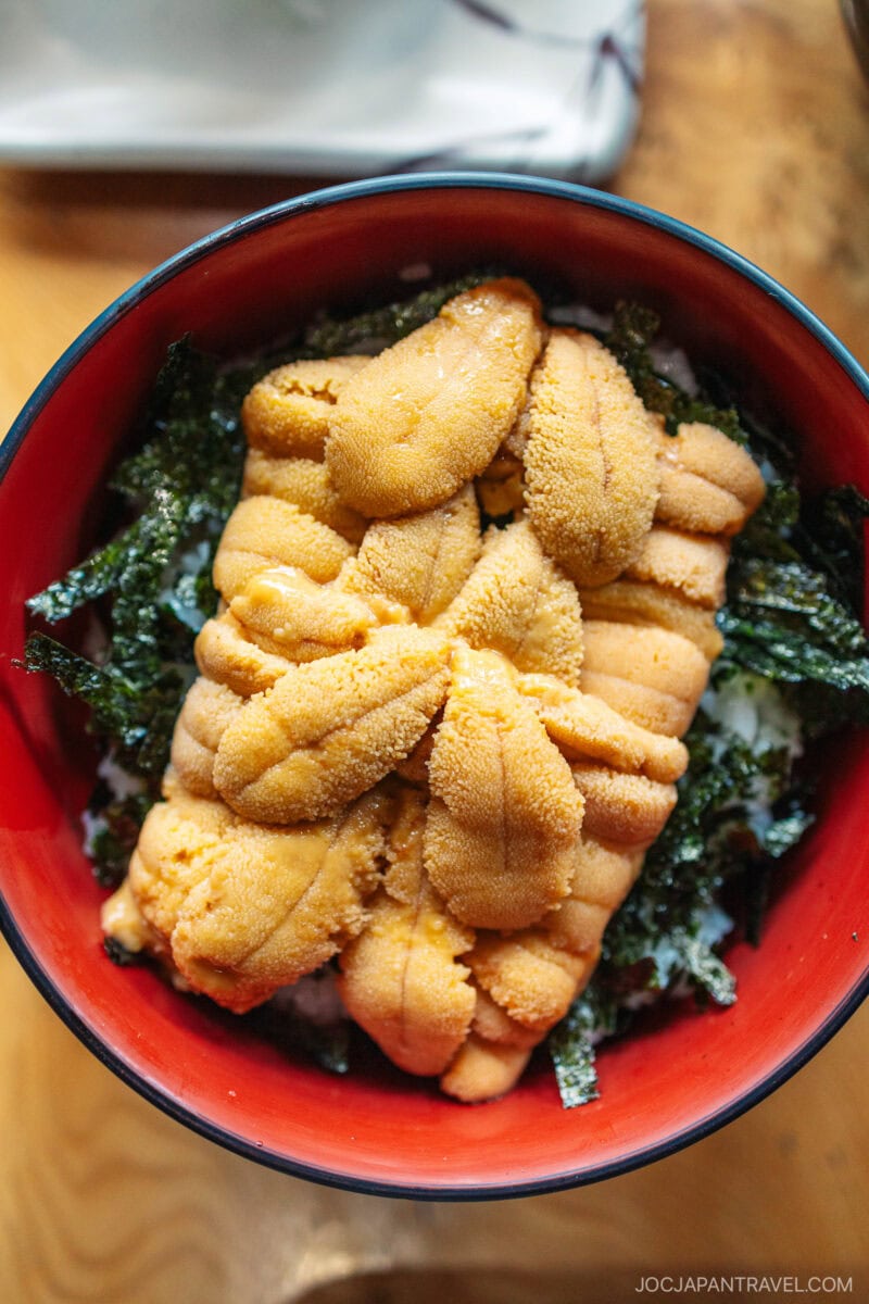A bowl of rice topped with fresh orange sea urchin (uni) and strips of green seaweed, served in a red and black bowl on a wooden table—a must-try dish from any Hokkaido food guide.