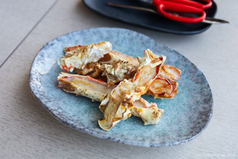A blue ceramic plate with several pieces of cooked crab legs, a highlight often featured in any Hokkaido food guide, sits on a light wooden table. In the background, a black tray holds a pair of red-handled scissors.