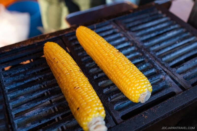 Two ears of corn on the cob grilling on a barbecue grill, with light charring visible on the kernels—perfect for any Hokkaido food guide seeking authentic local flavors.