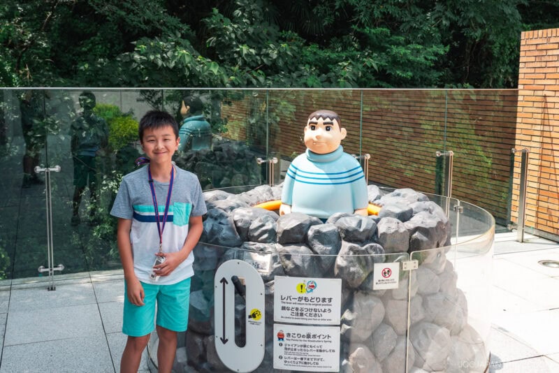 A smiling boy stands next to a cartoonish statue of a boy in a striped shirt emerging from faux rocks at the Fujiko F. Fujio Museum, with trees and a glass barrier in the background.
