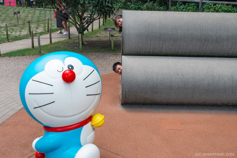 A large Doraemon statue stands in the foreground, while two people playfully peek their heads out from behind a cylindrical concrete structure in the background, capturing a fun moment at the Fujiko F. Fujio Museum park.