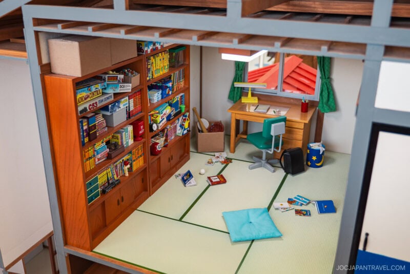 A tidy Japanese-style room with tatami mats, a bookshelf full of books and games, and floor cushions evokes the charm of the Fujiko F. Fujio Museum. Scattered manga and snacks add a playful touch; a blue backpack rests near the desk by the window.