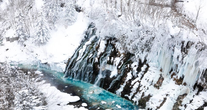 A partially frozen waterfall cascades down a rocky cliff surrounded by snow-covered trees, as featured in the Furano Biei winter guide, with turquoise water flowing below and icicles hanging from the rocks.