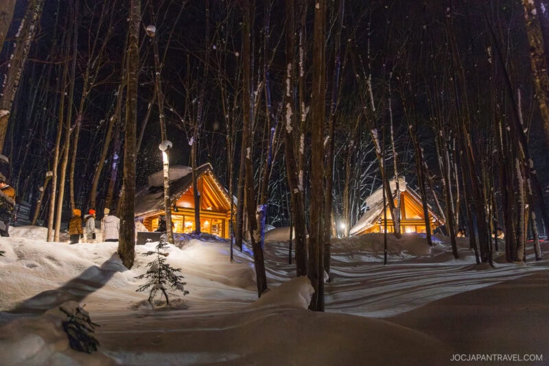 Log cabins glowing with warm light are nestled among tall, snow-covered trees at night, as people walk in the snowy forest—a cozy scene straight from a Furano Biei winter guide.