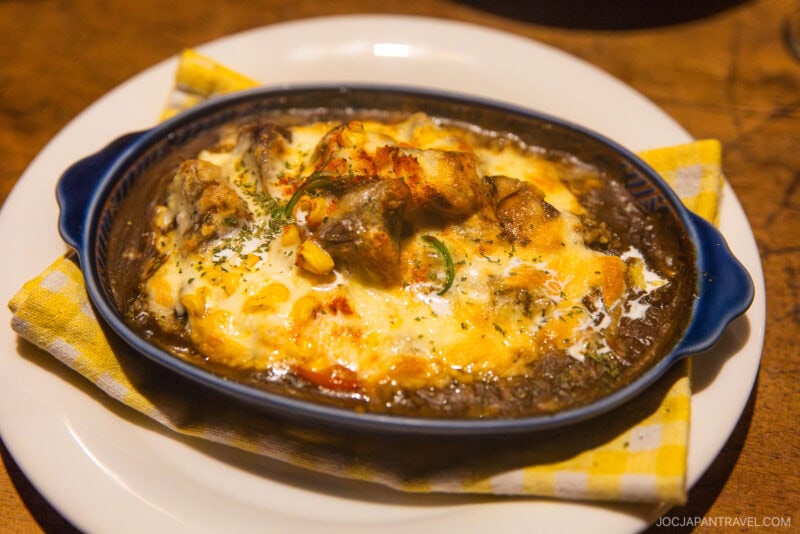 A baked dish of Japanese curry doria topped with melted cheese and herbs, served in a blue oval baking dish on a white plate with a yellow checkered napkin—perfect comfort food after following the Furano Biei winter guide.