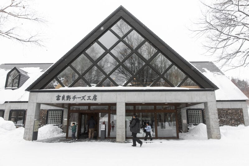 A modern building with a large glass facade and triangular roof, surrounded by snow. People walk near the entrance, while snow-covered trees create a scenic backdrop—an inviting scene from any Furano Biei winter guide.