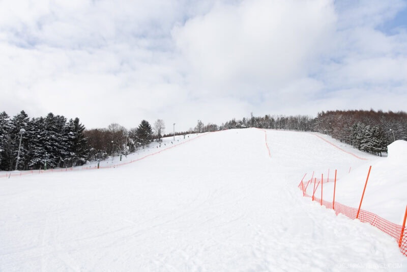 A snow-covered ski slope with orange safety netting on both sides, surrounded by trees under a partly cloudy sky—an iconic scene from the Furano Biei winter guide. No people are visible on the slope.